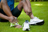 man sitting on the grass with one of his tennis shoes off as he ices his ankle with an ice pack.