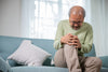 senior man sitting on his couch, holding his knee in pain.